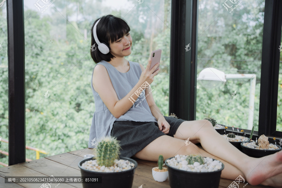 美丽的泰国亚裔女子在当地的仙人掌农场招待所用无线耳机和智能手机欣赏音乐。