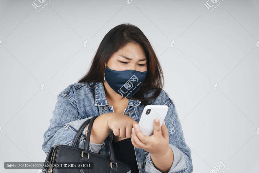 健康的亚洲胖女人戴着面具，穿着牛仔裤夹克，在白色背景下使用智能手机进行网上购物。