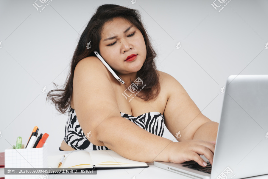 忙碌的亚洲胖乎乎的女人拿着笔记本电脑和电话，严肃地看着白色背景。