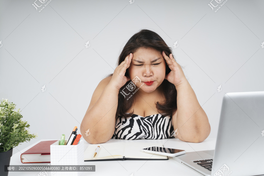 紧张的亚洲胖女人坐在办公桌前工作时头痛。