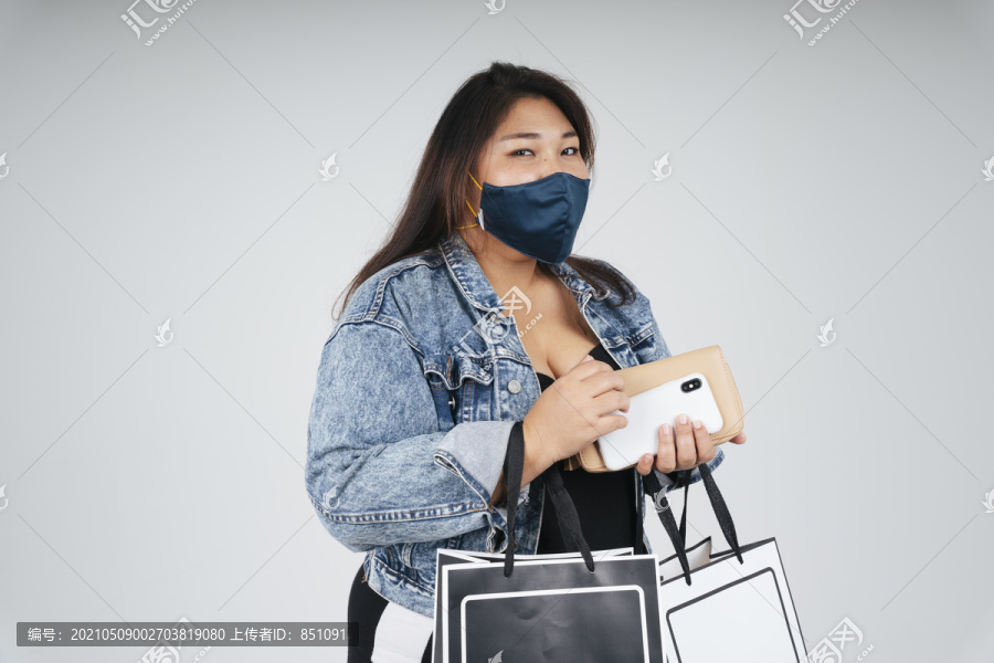 一个穿着黑色连衣裙和牛仔裤夹克的胖女人戴着面具，拿着纸袋，在白色的背景下。