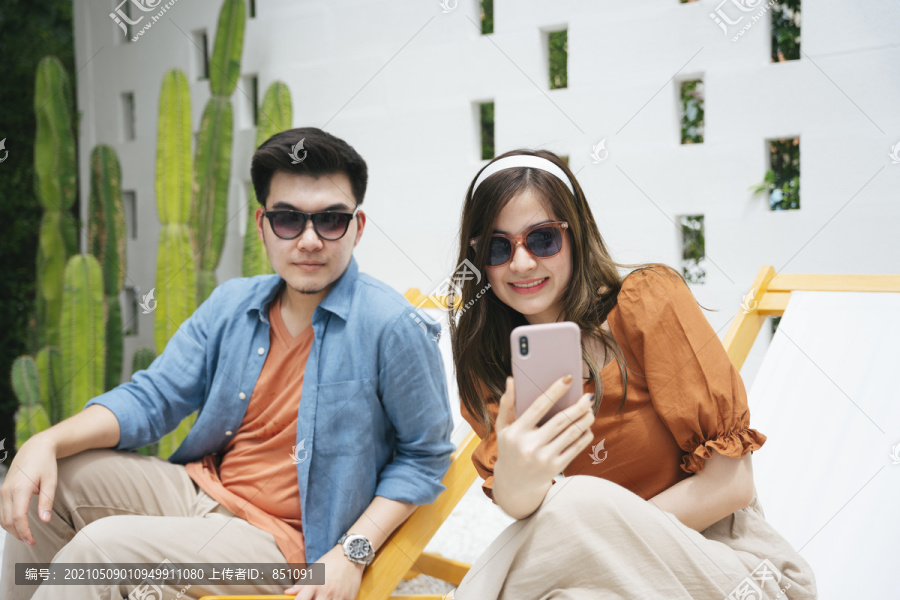 一对亚洲情侣戴着墨镜坐在沙滩上用智能手机自拍。