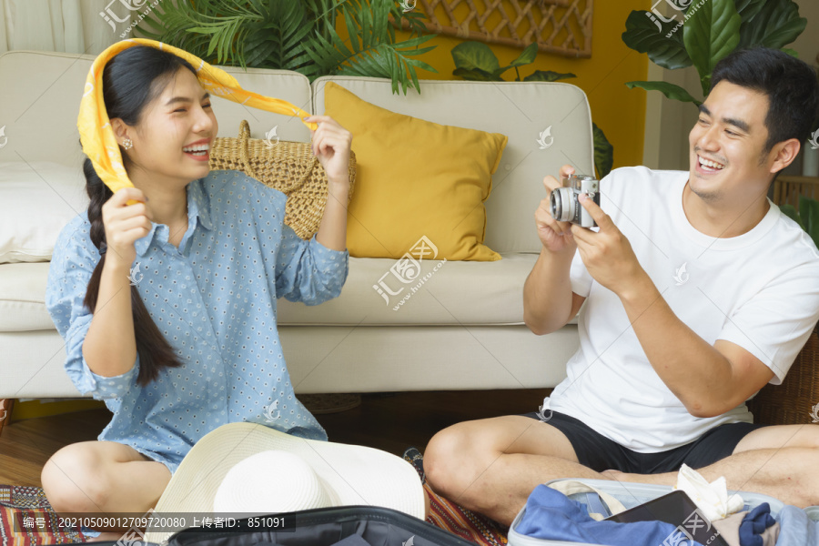 一对亚泰夫妇在酒店房间收拾东西时玩得很开心。男人用相机给妻子拍照。一个女人在被镜头拍下来的时候笑了。