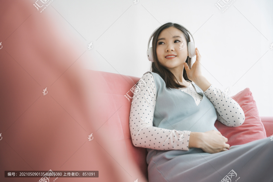 年轻漂亮的亚洲孕妇在沙发上用无线耳机听歌曲。