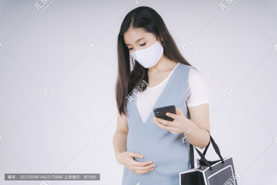 美丽的亚洲孕妇戴着棉质口罩用智能手机隔离白色背景。购物理念。