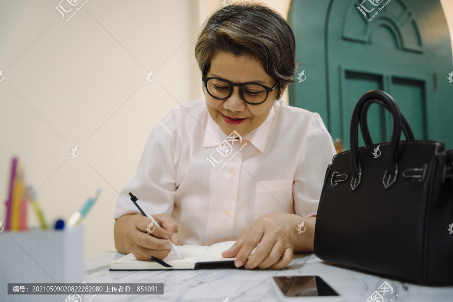 亚洲老年妇女戴眼镜在书上写日记。