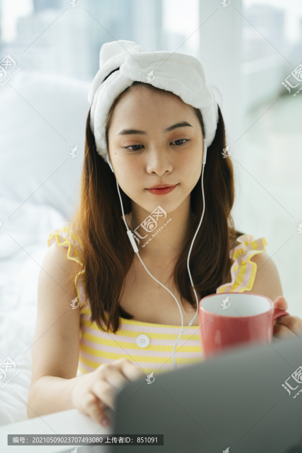 年轻的亚洲黑发女子戴着白色的头巾拿着一杯咖啡用耳机听音乐。