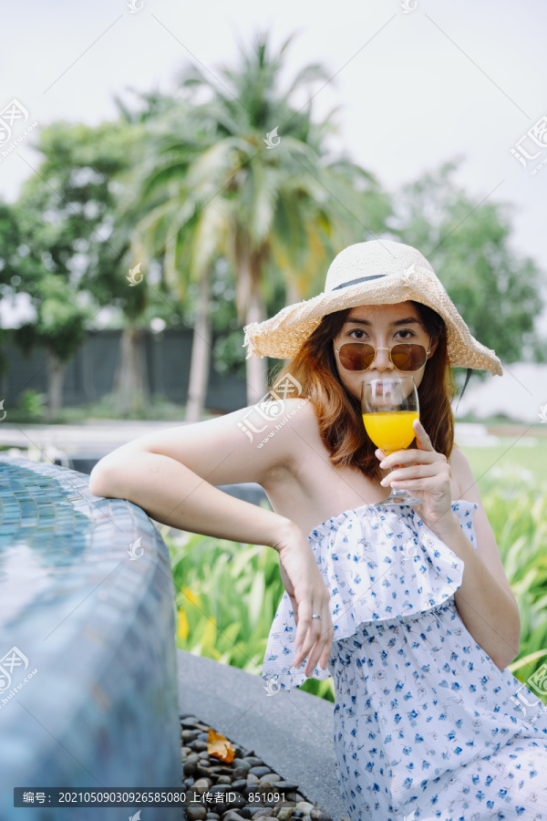 美丽的亚洲女游客戴着帽子和太阳镜在泳池边享受橙汁。