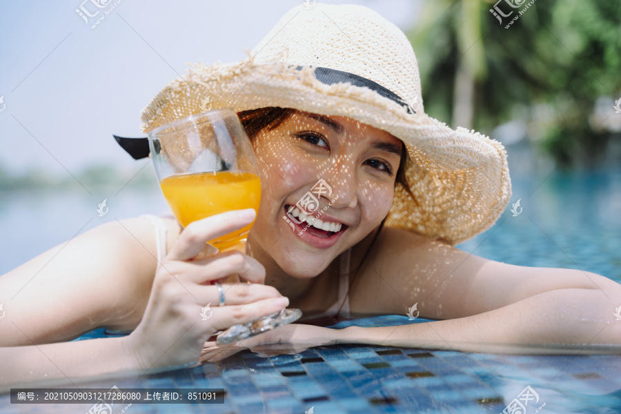 美丽的亚洲女游客戴着帽子捧着一杯橙汁在游泳池里放松。