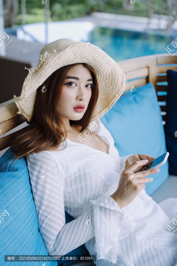 美丽的亚洲女游客戴着帽子手持智能手机在酒店休息室游泳池附近。