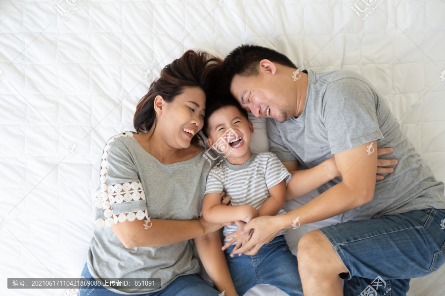 俯视图：可爱的小男孩和他的亚洲父母躺在家里的白色床上，看着相机，微笑着，幸福的家庭观念