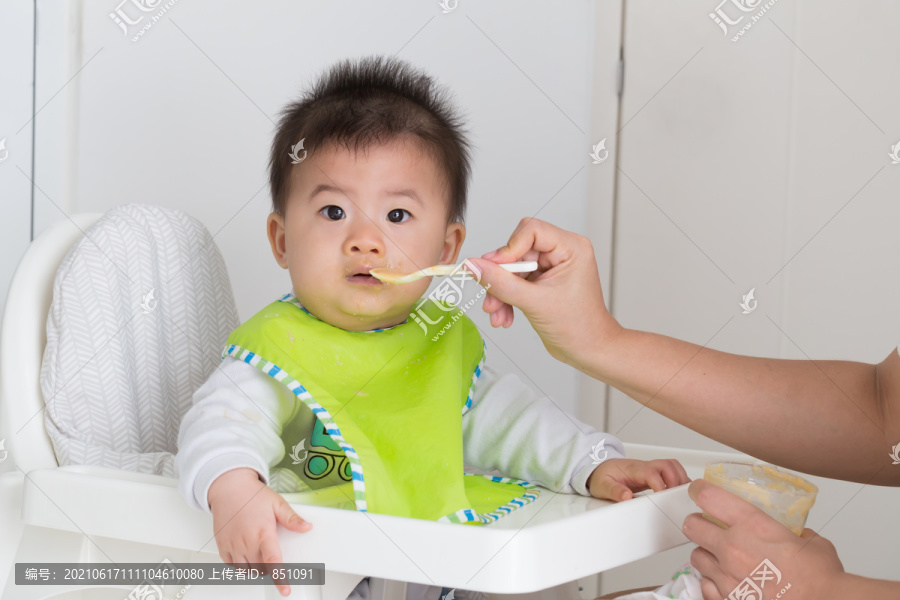 快乐的孩子坐在椅子上，妈妈在家里用勺子喂男孩，亚洲模特