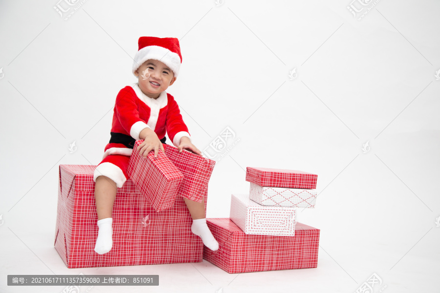 穿着圣诞老人套装的快乐亚洲男孩手持礼品盒，坐在白色背景上的红色礼品盒上