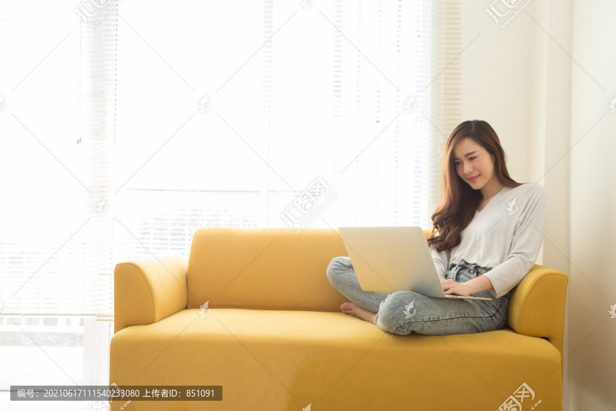 年轻的亚洲女人坐在家里办公室的黄色沙发上，手里拿着笔记本电脑。远程工作和在家工作的概念