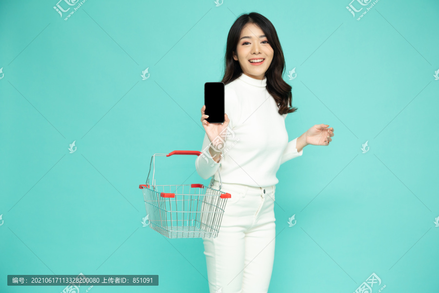 微笑着的快乐的亚洲女人拿着购物篮，在浅绿色的背景上展示手机