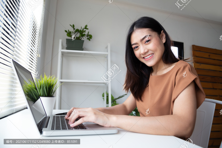 穿着休闲服的年轻亚洲女性在客厅室内工作时，使用笔记本电脑查看电子邮件，面带微笑。在家工作还是呆在家里