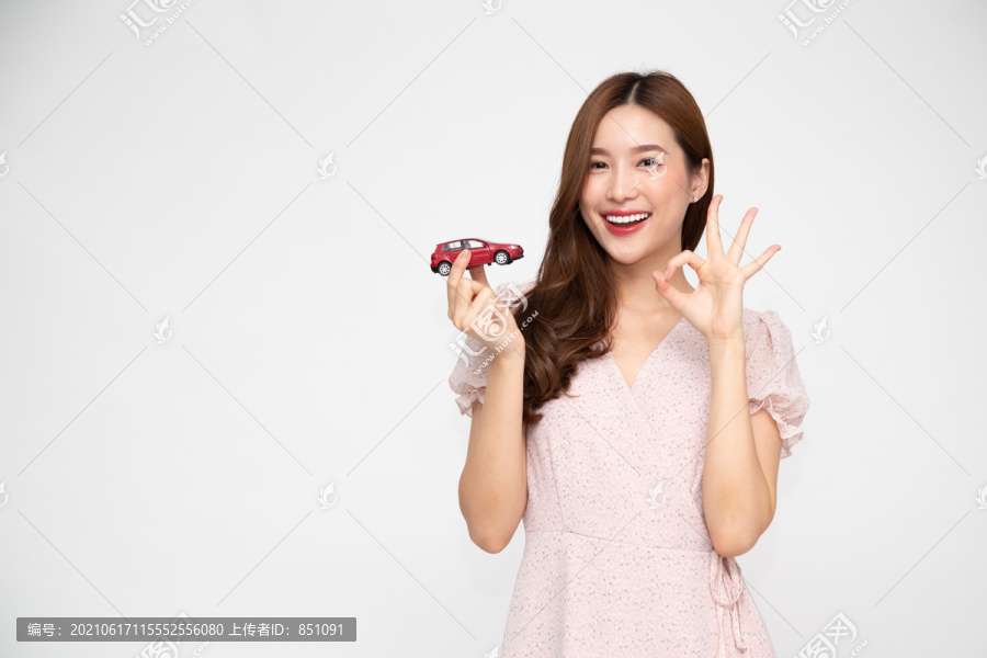 年轻的亚洲女性手持红色汽车模型，在白色背景上显示ok标志，旅游保险理念