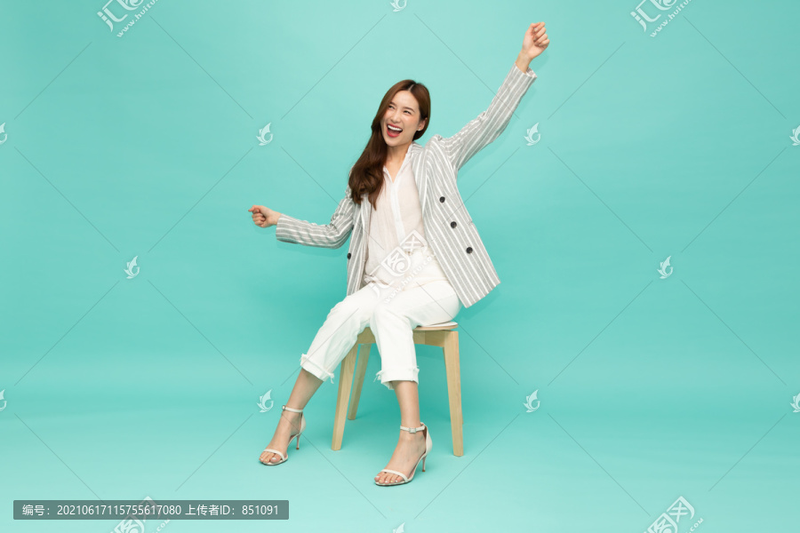 美丽迷人的亚洲女性坐在白色的现代椅子上，双手高举着幸福的双臂，兴奋的女商人成功的概念