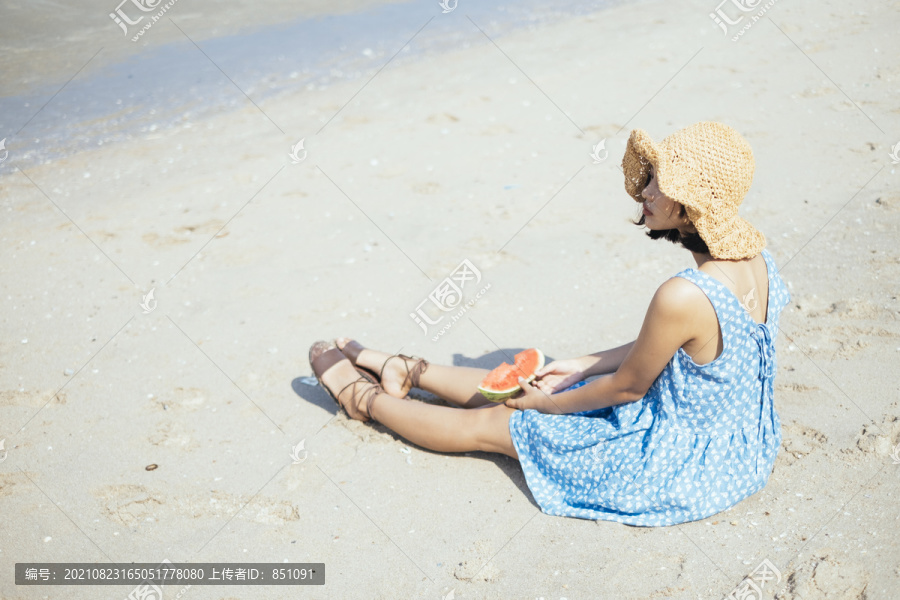可爱的黑发女孩坐在沙滩上，手里拿着一片西瓜，看着大海。