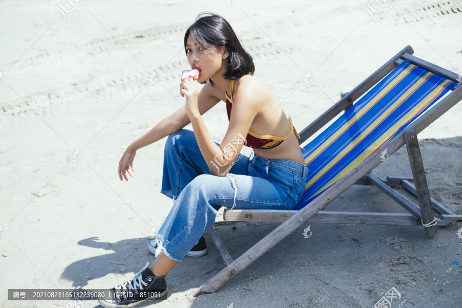穿着红色上衣的性感女孩在沙滩长椅上吃粉色蝴蝶冰淇淋。