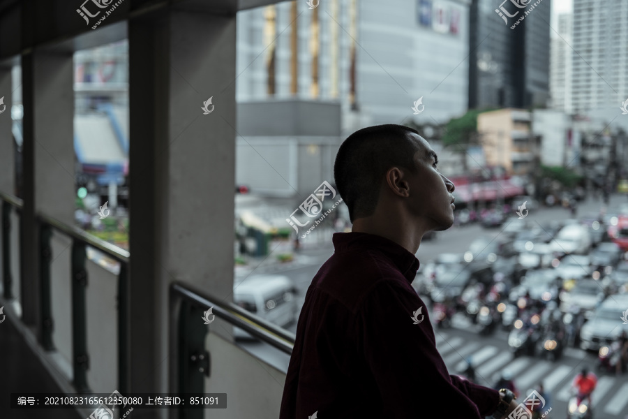 身穿红衫的光头男子站在满是汽车和摩托车的道路上方的天桥上。