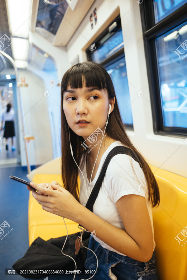 可爱的泰国年轻女子留着刘海，戴着耳机听音乐，坐在黄色的长椅上，坐在空中火车上。