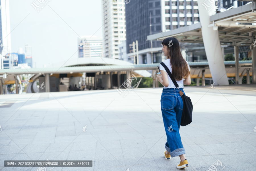 穿着白色衬衫和蓝色牛仔裤的年轻亚洲女人在城市里散步。城市旅游概念。