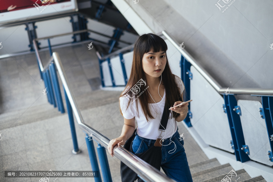 穿着白色衬衫和蓝色牛仔裤的可爱泰国年轻女子使用智能手机走上楼梯。