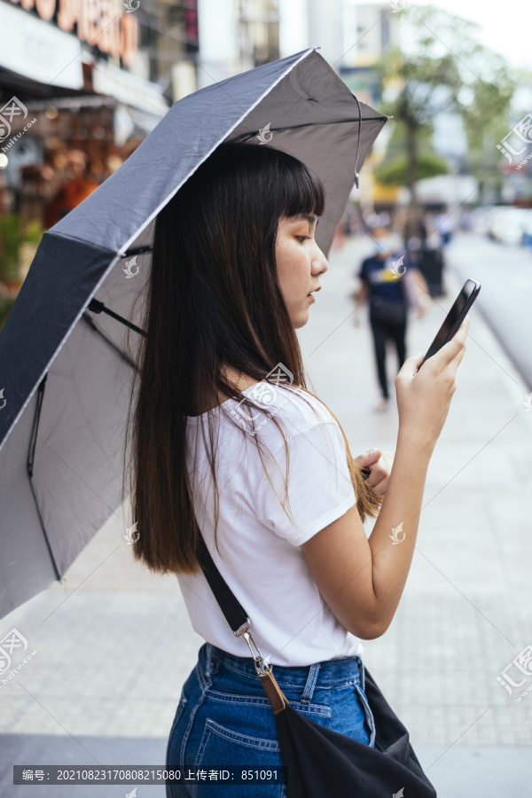 美丽的泰国年轻女子手持智能手机和雨伞，走在街上。