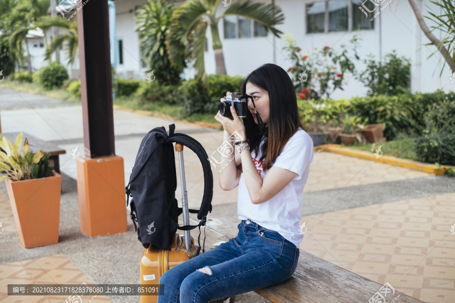 一位年轻的亚裔泰国长毛黑发女旅行者坐在长凳上等火车，手里拿着相机。