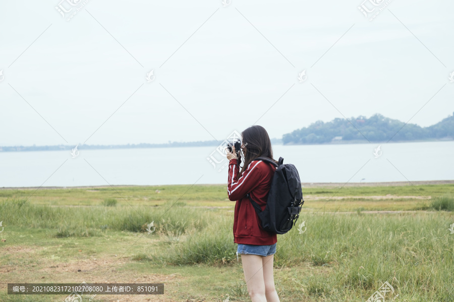 在春武里的湖边，一位年轻的亚裔泰国长发旅行者，身穿红色夹克，手持相机。