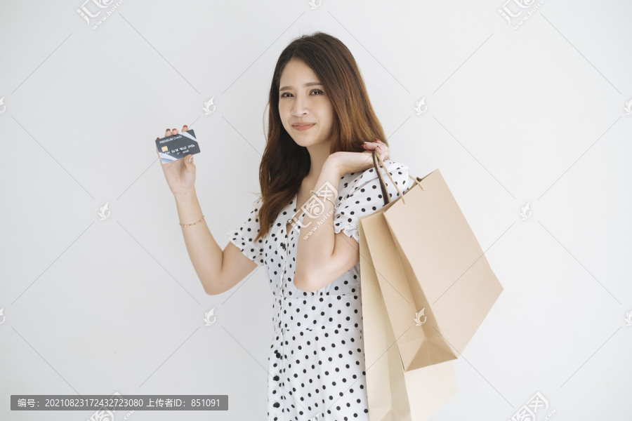 一位穿着黑白圆点连衣裙、手持信用卡、拎着购物纸袋、与世隔绝、背景为白色的快乐泰国妇女。网上购物的概念
