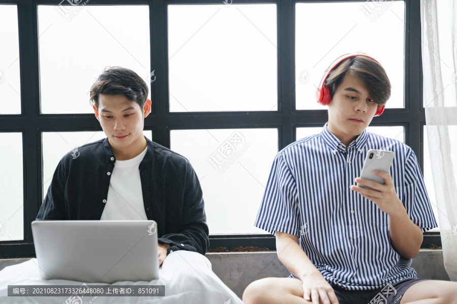 一对亚洲LGBTQ+男人住在房子里，用数字笔记本电脑工作，用耳机和智能手机听音乐。