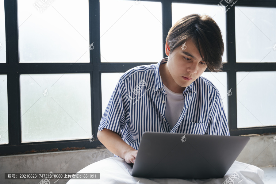 穿着休闲装的亚洲年轻人在家窗户附近的笔记本电脑上工作