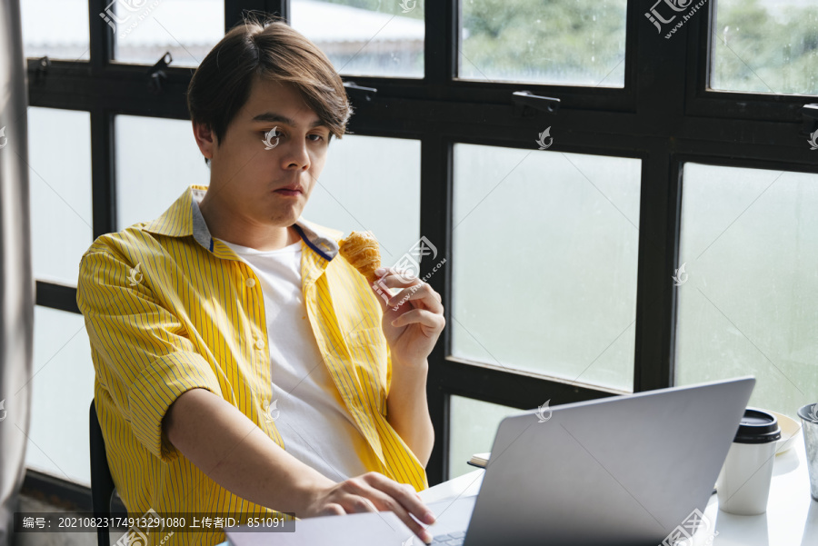 穿着黄衬衫的亚洲年轻人在现代办公室工作和吃零食。