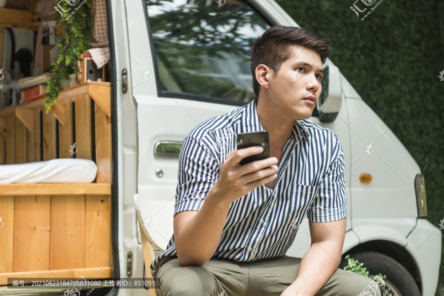 一名亚洲男子坐在停车场附近的椅子上，使用智能手机。