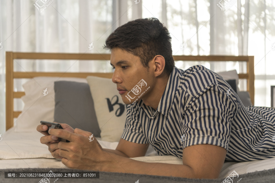亚洲男人躺在床上，在卧室里用智能手机玩手机游戏。