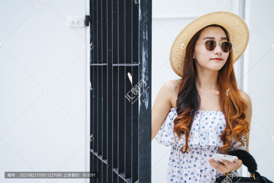 美丽的亚洲女旅行者戴着帽子和太阳镜，手里拿着智能手机站在门口。