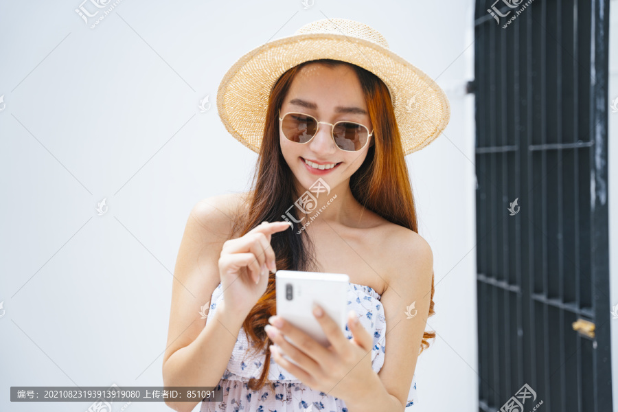 美丽的亚洲女旅行者戴着太阳镜，戴着帽子，在户外使用智能手机。