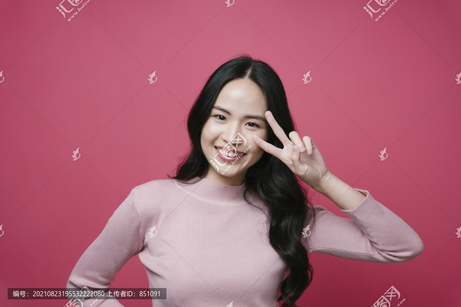 一位年轻的亚洲女性的肖像，在粉红色背景上展示了两个手指的胜利姿势。