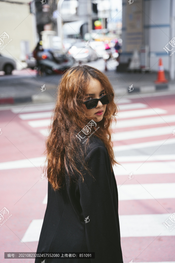 穿着墨镜和黑色夹克站在十字路口的年轻亚洲女性的时尚肖像。