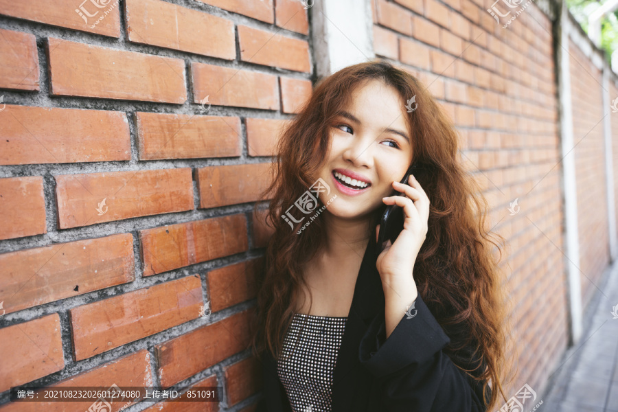 穿着黑色夹克的年轻亚洲女性在砖墙边打电话的时尚肖像。