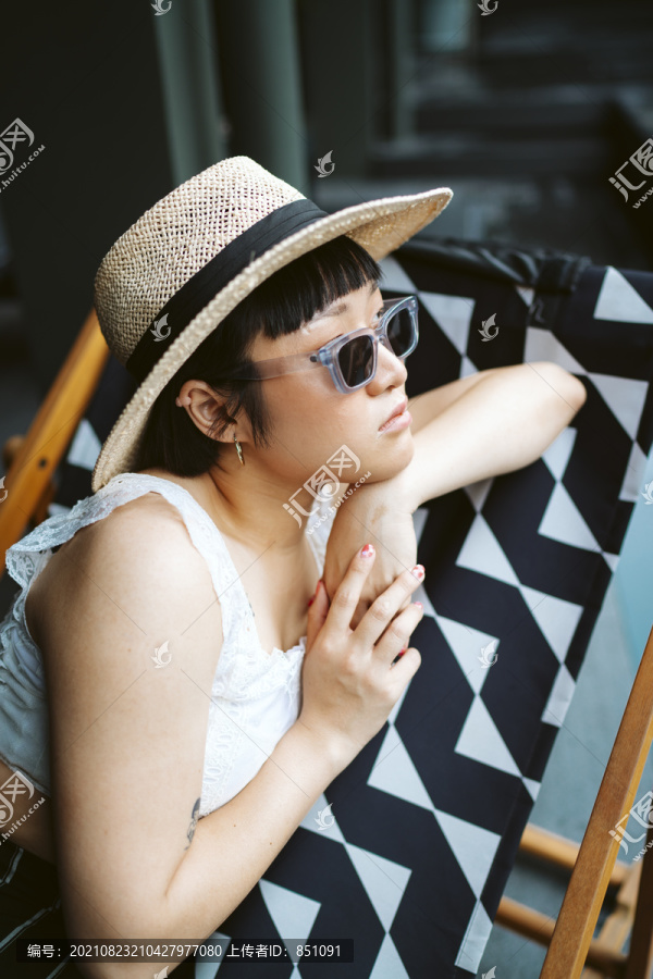 年轻的亚洲女人戴着太阳镜和帽子躺在酒店的沙滩椅上。