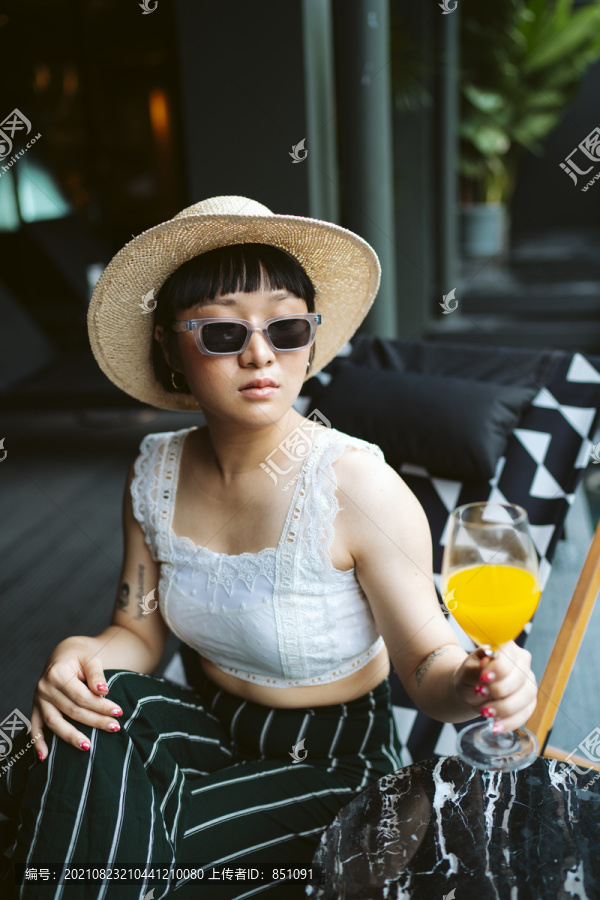一位戴着太阳镜和帽子在酒店喝橙汁的亚洲年轻女子。