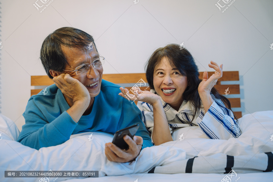 快乐的亚洲老年夫妇一起在卧室看电视。他们用智能手机控制面板。