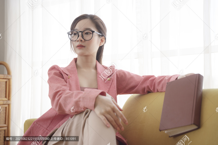 年轻的亚洲美女戴着眼镜，手里拿着一本书坐在沙发上。