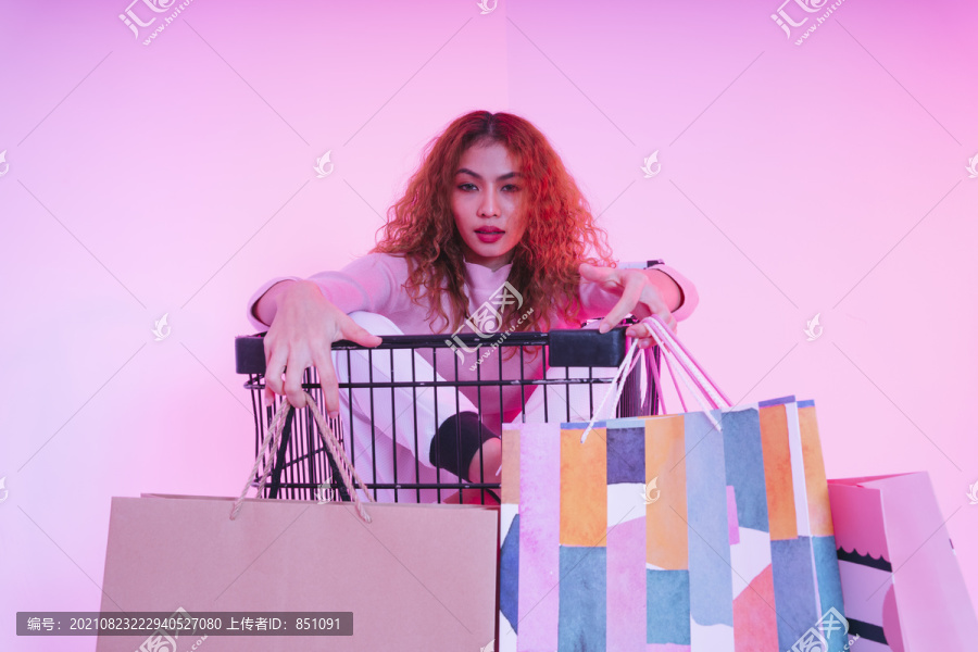 年轻的亚洲卷发女孩拿着纸袋坐在购物车上的肖像。