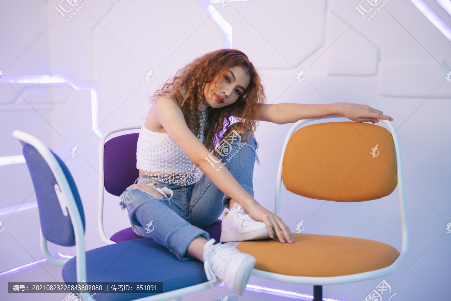 年轻的亚洲卷发美女的时尚肖像，穿着性感的白衬衫和牛仔裤裤子，坐在椅子上。