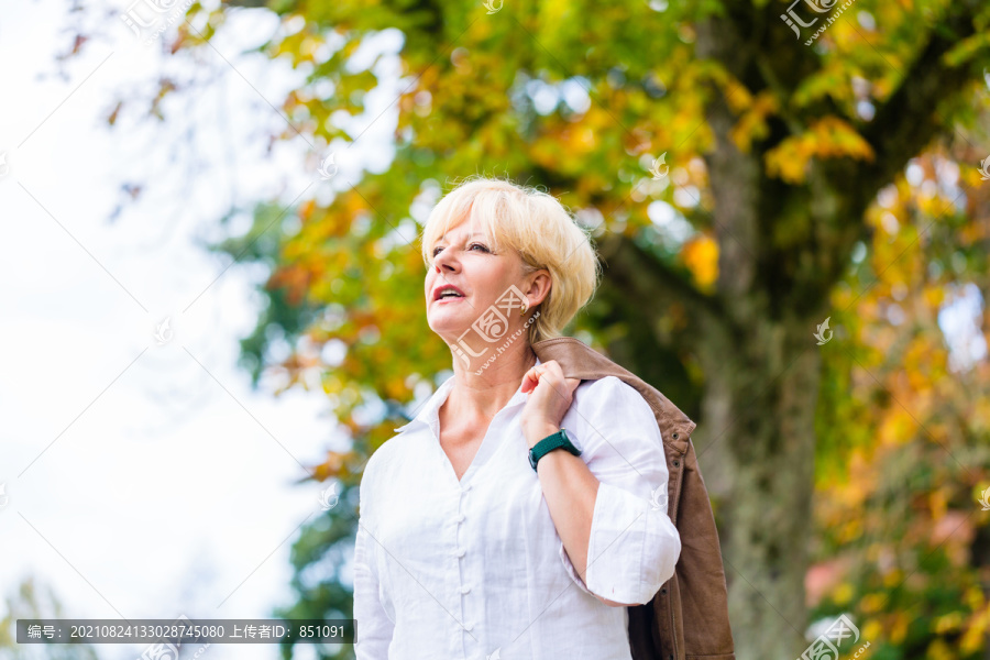 一位年长的女士在秋天的公园里走在五颜六色的树前，她的夹克衫搭在肩上