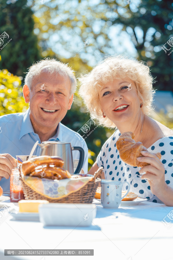 夏天，快乐的老人和女人坐在花园里吃早餐，吃面包卷，喝咖啡